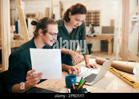 Zwei junge Zimmerleute, die in ihrer Werkstatt zusammen an einem Laptop arbeiten und planen, arbeiteten an etwas bedeutendem für unser Geschäft. Stockfoto