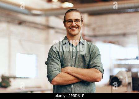 Möbel zu machen ist meine Art von Kunst. Porträt eines jungen Zimmermanns, der mit gefalteten Armen in einer Werkstatt posiert. Stockfoto