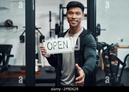 Ich heiße Sie in unserem Fitnesscenter willkommen. Verkürztes Porträt eines gutaussehenden jungen Fitnesstrainers, der seine Hand für einen Händedruck ausstreckt, während er ein Schild mit der Aufschrift „OFFEN“ in einem Fitnessstudio hochhält. Stockfoto