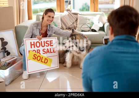 Waren die stolzen Besitzer jetzt. Ein glückliches junges Paar und ihr Hund, der Fotos mit einem verkauften Schild in ihrem neuen Zuhause macht. Stockfoto