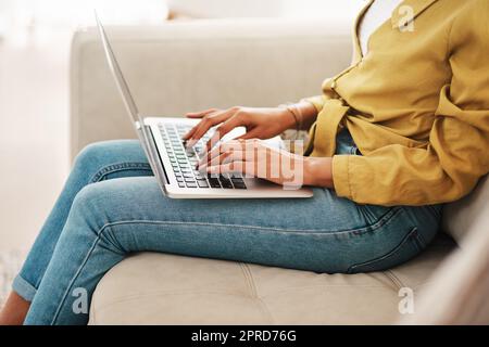 Ich bin immer am Erstellen. Eine unkenntliche Geschäftsfrau sitzt auf ihrer Couch und bloggt von ihrem Laptop, während zu Hause. Stockfoto