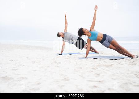 Gemeinsam einen aktiveren Lebensstil führen. Ein junger Mann und eine junge Frau üben gemeinsam am Strand Yoga. Stockfoto