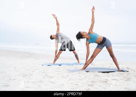 Yoga steht im Mittelpunkt ihres Wohlbefindens. Ein junger Mann und eine junge Frau praktizieren gemeinsam am Strand Yoga. Stockfoto