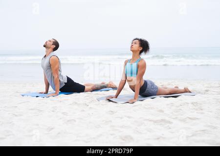 Lassen Sie die ruhige Luft Ihren Körper und Geist erfüllen. Ein junger Mann und eine junge Frau üben gemeinsam am Strand Yoga. Stockfoto