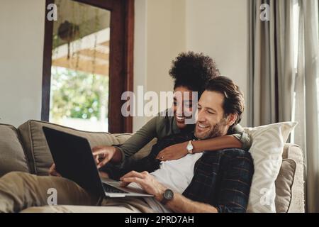 Schauen Sie sich diese Hüne an. Ein glückliches junges Paar, das einen Laptop benutzt, während es sich auf einer Couch im Wohnzimmer zu Hause entspannt. Stockfoto