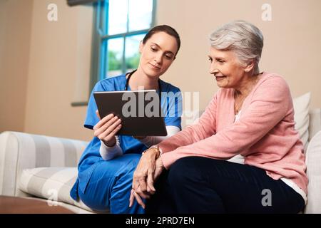 Ich wollte etwas schönes für Sie tun... eine Krankenschwester, die ihrer älteren Patientin etwas auf einem digitalen Tablet zeigt. Stockfoto