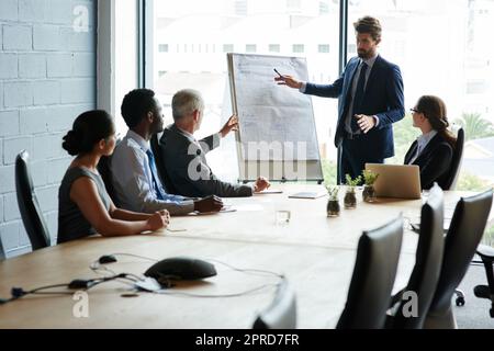 Selbstbewusster Geschäftsmann, der eine Präsentation auf einem Whiteboard mit verschiedenen Kollegen in einem Meeting in einer Vorstandssitzung führt. Manager verweist auf Forschungs- und Marketingdaten und plant gemeinsam mit seinem Team eine Strategie Stockfoto