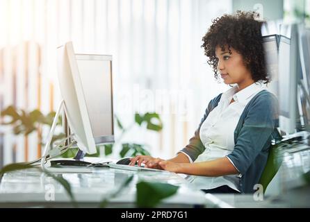 Junge Designerin, die an ihrem Computer an einem Schreibtisch in einem modernen Büro arbeitet. Kreative Geschäftsfrau, die am Arbeitsplatz Werbe- und Marketingdesigns auf ihre pc-Tastatur schreibt Stockfoto