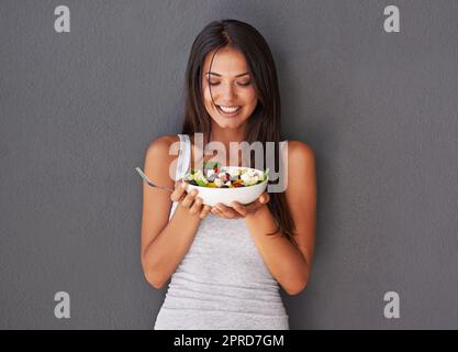 Eine gesunde junge Frau, die ihre frische Salatschüssel isst. Lächelnde, wunderschöne Frau, die im Rahmen ihres veganen Wellness-Lebensstils ihre saubere, grüne Diät mit Gemüse hält und genießt Stockfoto