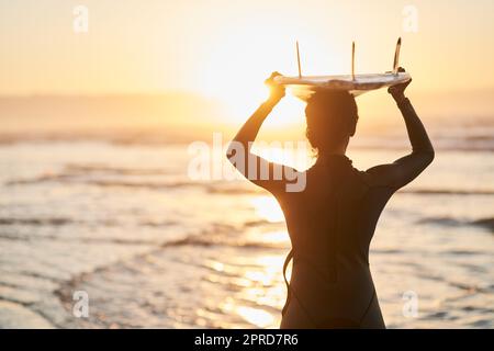 Ich bin genau dort, wo Ive immer hingehörte. Rückansicht einer Surferin, die ihr Surfbrett am Strand über ihren Kopf trägt. Stockfoto