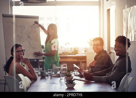 Glückliche Geschäftsfrau, die an Bord schreibt und Ideen und Gedanken erklärt, während sie mit Kollegen Brainstorming führt. Porträt verschiedener Kollegen, die während der Zusammenarbeit an einem Projekt Ideen diskutieren Stockfoto