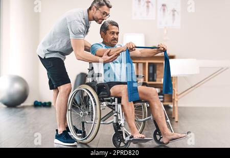 Stärke liegt im Gleichgewicht. Ganzkörperaufnahme eines älteren Mannes in einem Rollstuhl, der mit einem Widerstandsband neben seinem Physiotherapeuten trainiert. Stockfoto