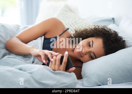 Eine attraktive junge Frau lag auf ihrer Seite im Bett und benutzte morgens ihr Handy. Stockfoto