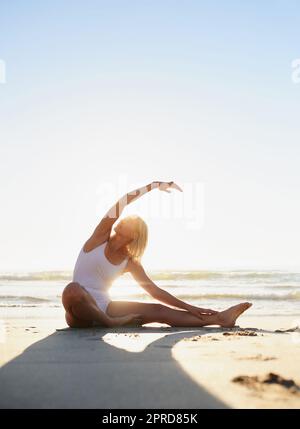 Achtet darauf, wie sich mein Körper anfühlt. In voller Länge eine attraktive junge Frau, die am frühen Morgen am Strand eine Yoga-Strecke macht. Stockfoto
