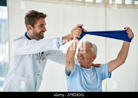 Ein freundlicher junger Arzt half einem älteren Mann mit einem Stretching Band in seinem Büro zu strecken. Stockfoto