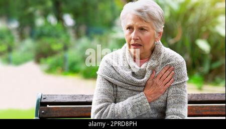 Ich muss den Arzt bald wieder aufsuchen. Eine gestresste ältere Frau sitzt auf einer Bank und hält ihre Brust in Unannehmlichkeiten draußen in einem Park. Stockfoto