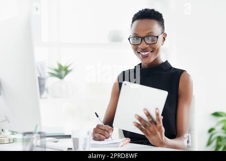 Es fühlt sich nicht an, als würde man das lieben, was man tut. Eine selbstbewusste junge Geschäftsfrau schreibt in einem Notebook und benutzt ein digitales Tablet in einem modernen Büro. Stockfoto