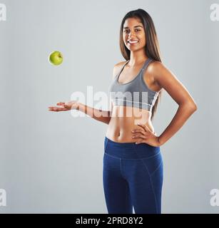 Ich mag es, ein wenig grün auf meine Ernährung. Beschnittenes Studioporträt einer glücklichen jungen Frau, die einen Apfel auf einen grauen Hintergrund wirft. Stockfoto