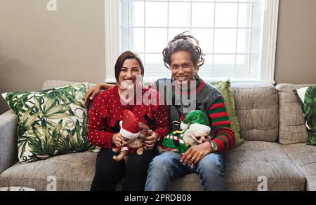 Unsere Hunde sind unsere anderen Kinder. Porträt eines fröhlichen jungen Paares, das seine Hunde hält, während es zu hause während der weihnachtszeit auf einem Sofa sitzt. Stockfoto