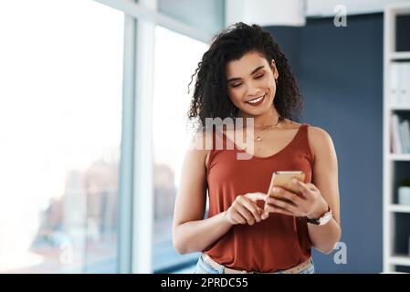 Antworten auf SMS, bevor sie wieder zur Arbeit kommen. Eine attraktive junge Geschäftsfrau, die allein in ihrem Büro steht und auf ihrem Handy schreibt. Stockfoto
