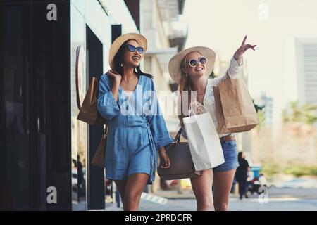 Wir sollten definitiv in den Laden gehen. Eine attraktive junge Frau, die auf etwas zeigt, während sie mit ihrer besten Freundin während einer Shopping-Tour in der Stadt spaziert. Stockfoto