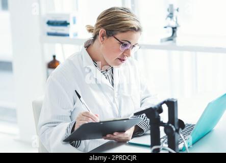 Das sieht aus wie etwas, das ich vorher gesehen habe: Eine attraktive junge Wissenschaftlerin, die ein Dokument ausfüllt, während sie einen Laptop in einem Labor benutzt. Stockfoto