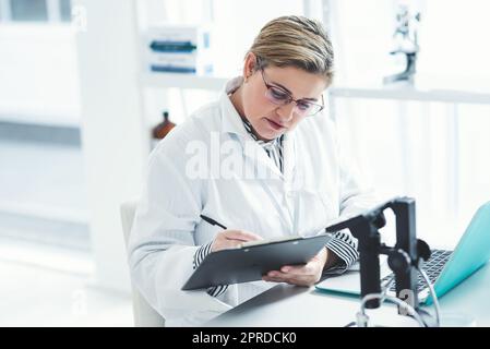 Eine attraktive junge Wissenschaftlerin, die ein Dokument ausfüllt, während sie einen Laptop in einem Labor benutzt. Stockfoto