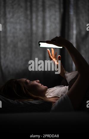 Junge Frau im Bett hält ein Telefon, müde und erschöpft, blaues Licht belastet ihre Augen, vermasseln ihren zirkadianen Rhythmus Stockfoto