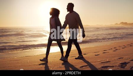 Gingen dorthin, wo die Liebe uns führt. Ganzkörperaufnahme eines liebevollen jungen Paares, das bei Sonnenuntergang am Strand spazieren geht. Stockfoto