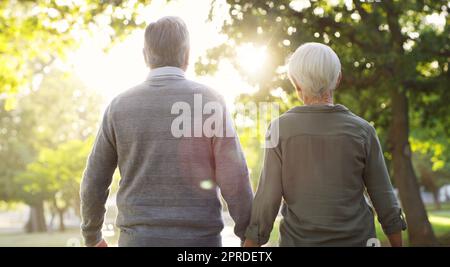 Liebe auf der anderen Seite der Jugend. Rückansicht eines liebevollen Seniorenpaares, das zusammen läuft und dabei die Hände im Park hält. Stockfoto