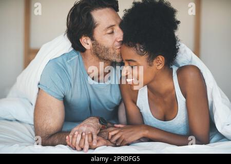 Ein glückliches, sorgloses und liebevolles Paar teilt sich morgens einen Kuss, der zusammen im Bett liegt. Gemischtrassig, intim und fürsorglich Ehemann und Ehefrau, verbunden und liebevoll, während sie sich zu Hause verbinden Stockfoto