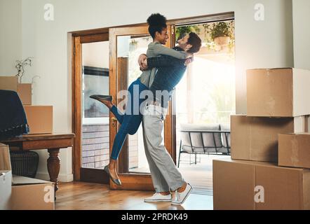 Ein freudiges, gemischtrassiges Paar zieht zusammen in ein neues Zuhause und umarmt sich, glücklich und aufgeregt. Vielfältige, liebevolle und junge Liebhaber ziehen in ein Haus um und feiern durch Umarmung Stockfoto