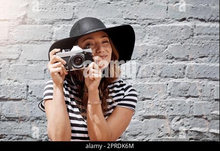 Jeden Moment in der Stadt festhalten. Eine junge Frau, die mit ihrer Kamera im Freien gegen eine Backsteinmauer fotografiert. Stockfoto