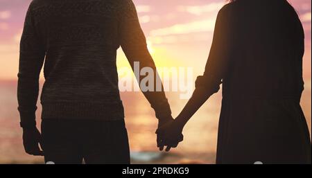 Wahre Liebe bei Sonnenuntergang. Rückansicht eines unerkennbaren, liebevollen Paares, das sich bei Sonnenuntergang am Strand die Hände hält. Stockfoto