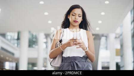 Unterwegs arbeiten. Eine junge Geschäftsfrau, die ein Smartphone benutzt, während sie durch ein modernes Büro geht. Stockfoto