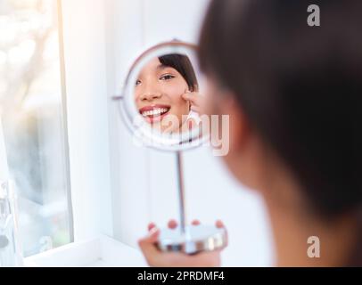 Eine fröhliche, attraktive junge Frau, die Feuchtigkeitscreme auf ihr Gesicht aufgibt, während sie im Spiegel in ihre Spiegelung schaut. Stockfoto