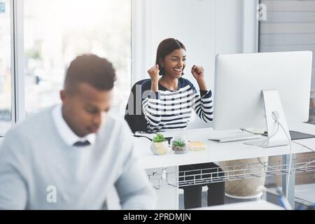 Selbst die kleinsten Siege verdienen es, gefeiert zu werden. Eine junge Geschäftsfrau feiert an ihrem Schreibtisch mit einem Kollegen, der im Vordergrund arbeitet. Stockfoto