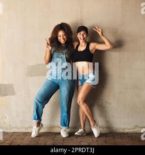 Wir kommen in Frieden. Porträt von zwei fröhlichen jungen Frauen, die sich tagsüber für ein Foto posieren, während sie sich draußen an einer Wand lehnen. Stockfoto