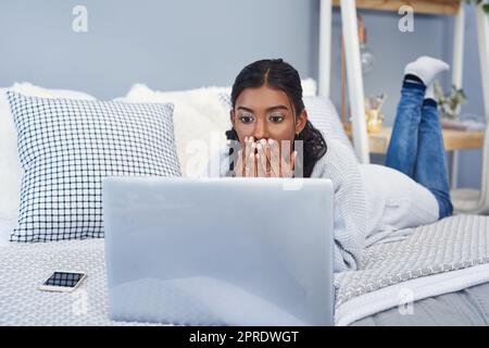 Sie kann nicht glauben, was sie online liest. Eine attraktive junge Frau, die einen Laptop benutzt, während sie sich in ihrem Schlafzimmer zu Hause auf ihrem Bett abkühlte. Stockfoto