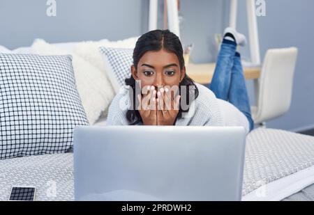 Ich kann nicht glauben, was online gesagt wird. Eine attraktive junge Frau, die einen Laptop benutzt, während sie sich in ihrem Schlafzimmer zu Hause auf ihrem Bett abkühlte. Stockfoto