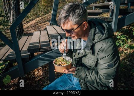 Ein Mann isst auf einem Salat aus einer Einwegpapierschüssel im Freien während eines Spaziergangs in der Natur im Herbst. Stockfoto