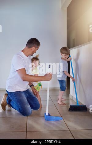 Teamarbeit macht es schneller und besser. Zwei kleine Geschwister helfen ihrem Vater zu Hause, den Boden zu kehren. Stockfoto