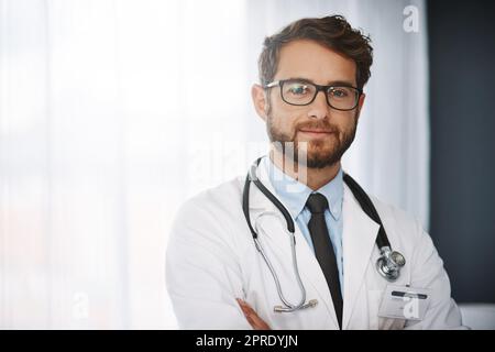 Ihre Gesundheit ist meine oberste Priorität. Beschnittenes Porträt eines selbstbewussten jungen männlichen Arztes, der mit gefalteten Armen in einem Krankenhaus steht. Stockfoto