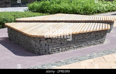 Holzbänke aus Gabionenkörben mit Steinen im Inneren. Holz auf Gabionen montiert. Bänke in modernem Design. Es gibt einen Erholungsbereich. Detail einer niedrigen Gabionenwand mit Holzplatte. Stockfoto