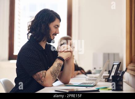 Junger, glücklicher und trendiger Geschäftsmann, der in einem Büro an seinem Laptop arbeitet. Ein Mann auf einem Online-Geschäftstreffen, der über sein Start-up spricht. Professioneller Typ, der am Schreibtisch sitzt und einen Sprachassistenten auf Abruf benutzt Stockfoto