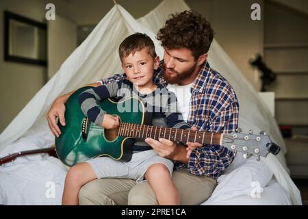 Ich lerne, Gitarre zu spielen. Ein beschnittenes Porträt eines entzückenden kleinen Jungen, der auf dem Schoß seines Vatis sitzt, während er zuhause Gitarre spielen lernt. Stockfoto