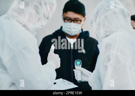 Negativer Covid-Test und Daumen hoch für jungen Mann aus dem Gesundheitswesen nach dem Test auf Virus. Medizinische Fachkräfte in Gefahrstoffanzügen, die Temperaturtests während des Ausbruchs oder der Pandemie machen Stockfoto