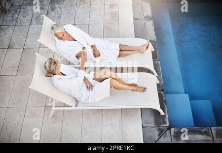 Die Sonne einholen. Aufnahmen aus einem hohen Winkel eines fröhlichen Paares mittleren Alters, das tagsüber in Bademänteln neben einem Swimmingpool im Spa im Freien entspannt. Stockfoto