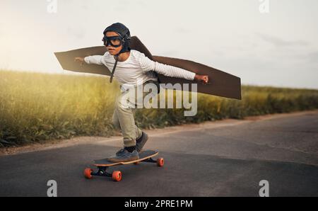 Ein kleiner Junge, der vorgibt, mit einem Paar Pappflügeln zu fliegen, während er draußen auf einem Skateboard reitet. Stockfoto