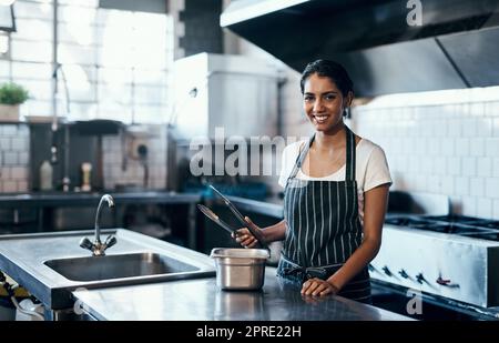 Kochen, Kochen und Arbeiten als Koch in einer kommerziellen Küche mit Zangen und industrieller Ausrüstung. Porträt einer weiblichen Köchin, die eine Mahlzeit zum Mittag-, Abendessen oder Abendessen in einem Restaurant oder Café zubereitet Stockfoto